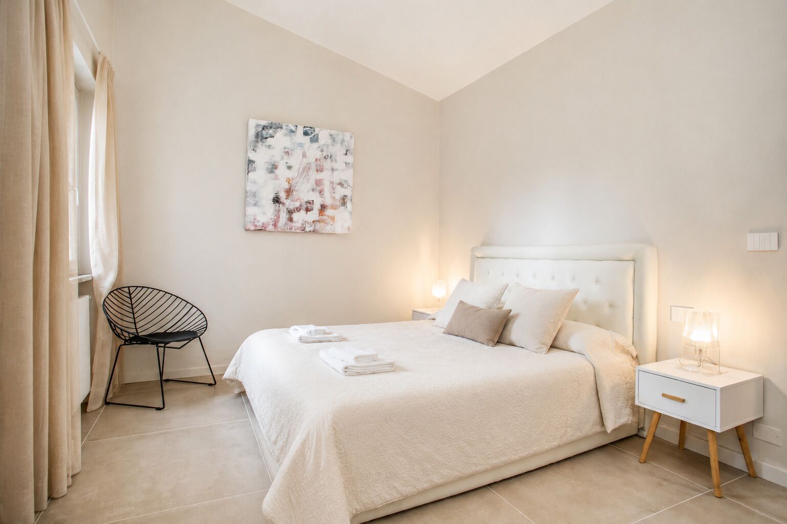 A modern bedroom with a large bed, a chair, a bedside table with a lamp, and a painting on the wall.