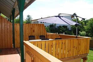 Une terrasse en bois avec une table et des chaises, un espace couvert et une vue sur un bâtiment en arrière-plan.