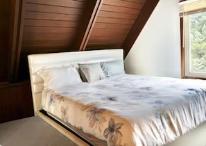 Un dormitorio con techo inclinado, una cama con ropa de cama floral y un ventanal con vistas a árboles.