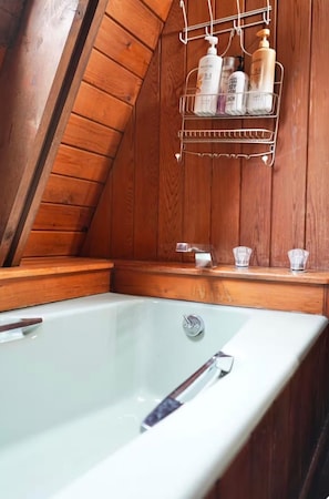 Interior de una cabaña de madera con una bañera, un estante metálico con artículos de aseo y un banco de madera.