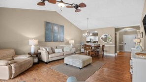 A spacious living room with a high ceiling, a dining area with a chandelier, and a kitchen visible through an archway. - 832 Cypress Bend - Barefoot Resort Rentals (North Myrtle Beach)