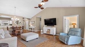 Ein geräumiges Wohnzimmer mit Fernseher, Deckenventilator und Essbereich mit einer Chandelier.