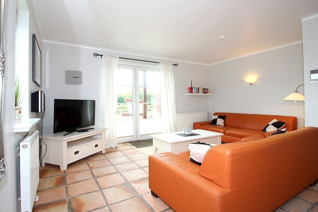 A living room with a television, orange sofa, white coffee table, and a window with curtains.