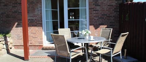An outdoor patio with a round table and four wicker chairs, a brick wall, and a sliding glass door.
