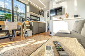 A modern living room with a sofa, a coffee table, and a TV. There is a kitchen area with a microwave and a coffee machine.