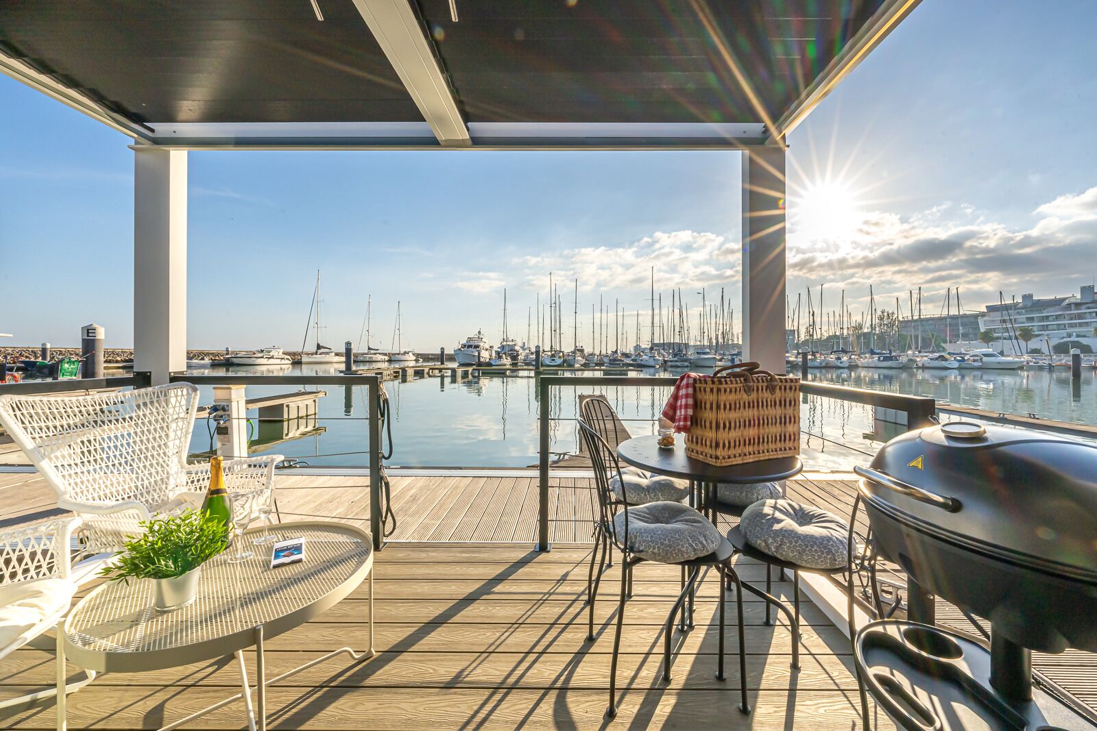 A covered outdoor area with a view of a marina, featuring a grill, seating, and a bottle of champagne.