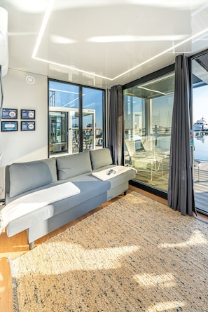 A modern living room with a grey sofa, a glass door leading outside, and a view of boats.