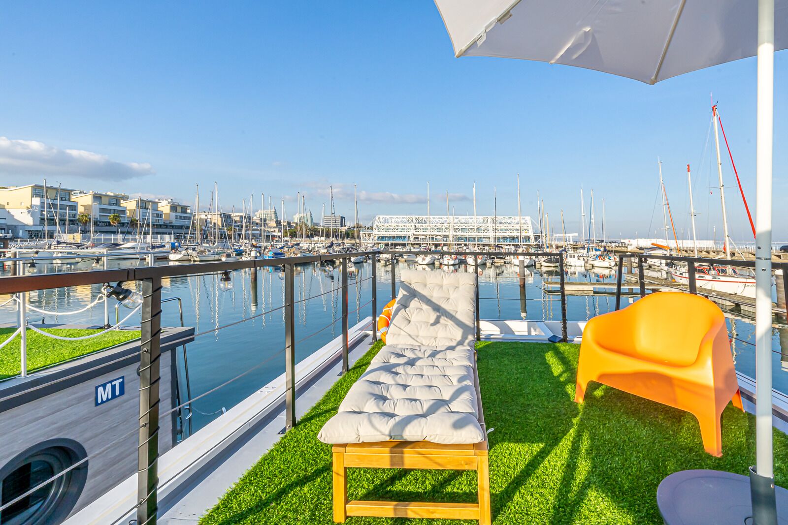 A balcony with a view of a marina, a lounge chair, a small table, and a white umbrella.