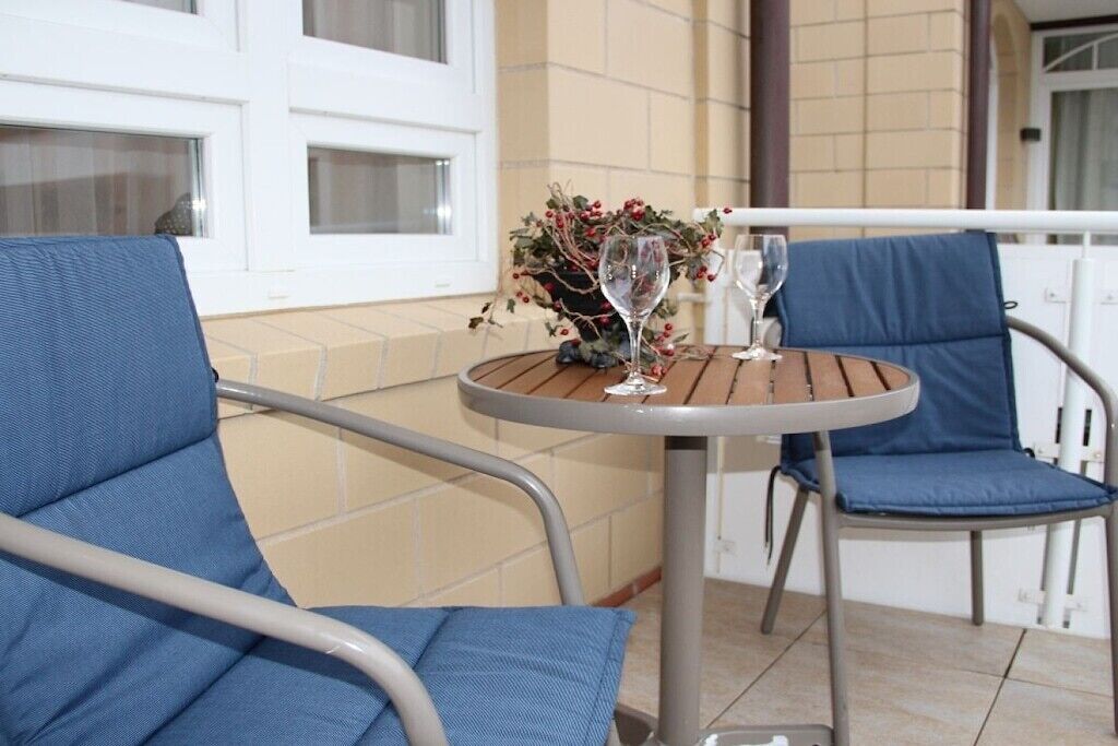 A round metal table with two chairs, a potted plant, and two wine glasses on a balcony with tiled flooring and white railings.