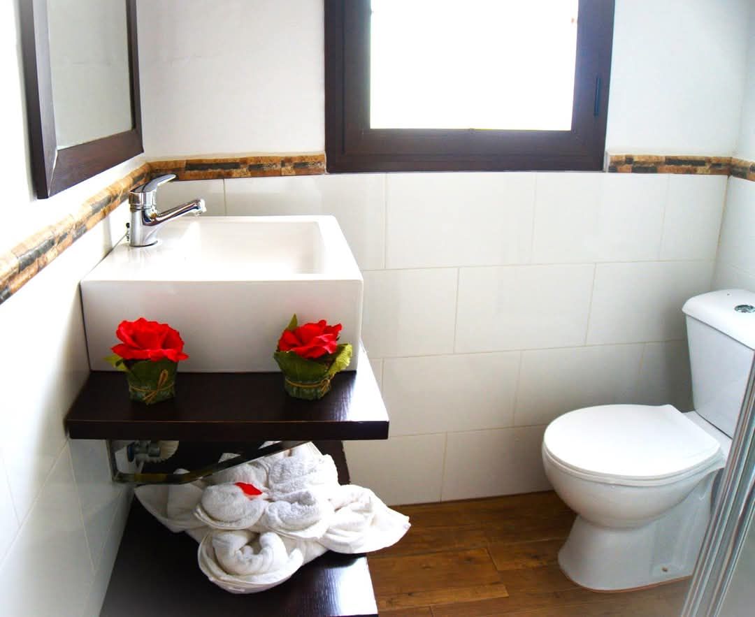 Un baño con un lavamanos blanco, dos rosas rojas, un inodoro y una ventana sobre el lavamanos.