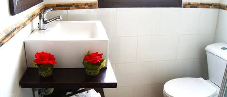 Un baño con un lavamanos blanco, dos rosas rojas, un inodoro y una ventana sobre el lavamanos.