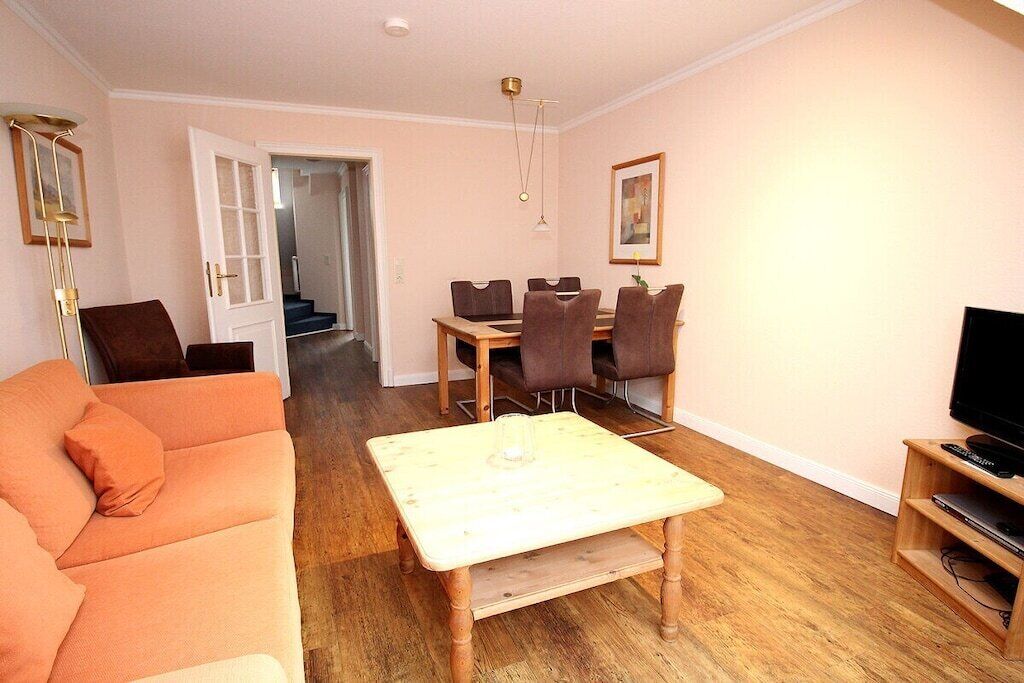 Una sala de estar con sofá, una mesa de centro, una mesa de comedor con sillas, un televisor y una escalera que conduce a otra habitación.