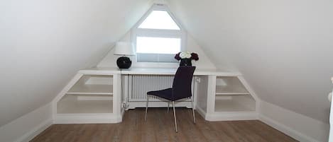 A room with a sloped ceiling, a window, a desk with a chair, and built-in shelving.