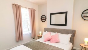 A hotel room with a bed, a bedside table with a lamp, a round mirror, and a window with curtains. - Regents Cottage (Melton Mowbray)
