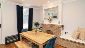A dining area with a wooden table, benches, a brick fireplace, a mirror above the mantel, and a window with curtains. - Regents Cottage (Melton Mowbray)