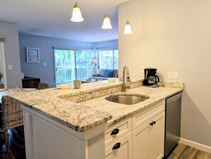 A kitchen with a granite countertop, stainless steel sink, and a coffee maker. There is a dining area with wicker chairs and a sofa. - Sea Colony 3BR Condo w/ Blue Screened Porch & Resort Amenities (Bethany Beach)