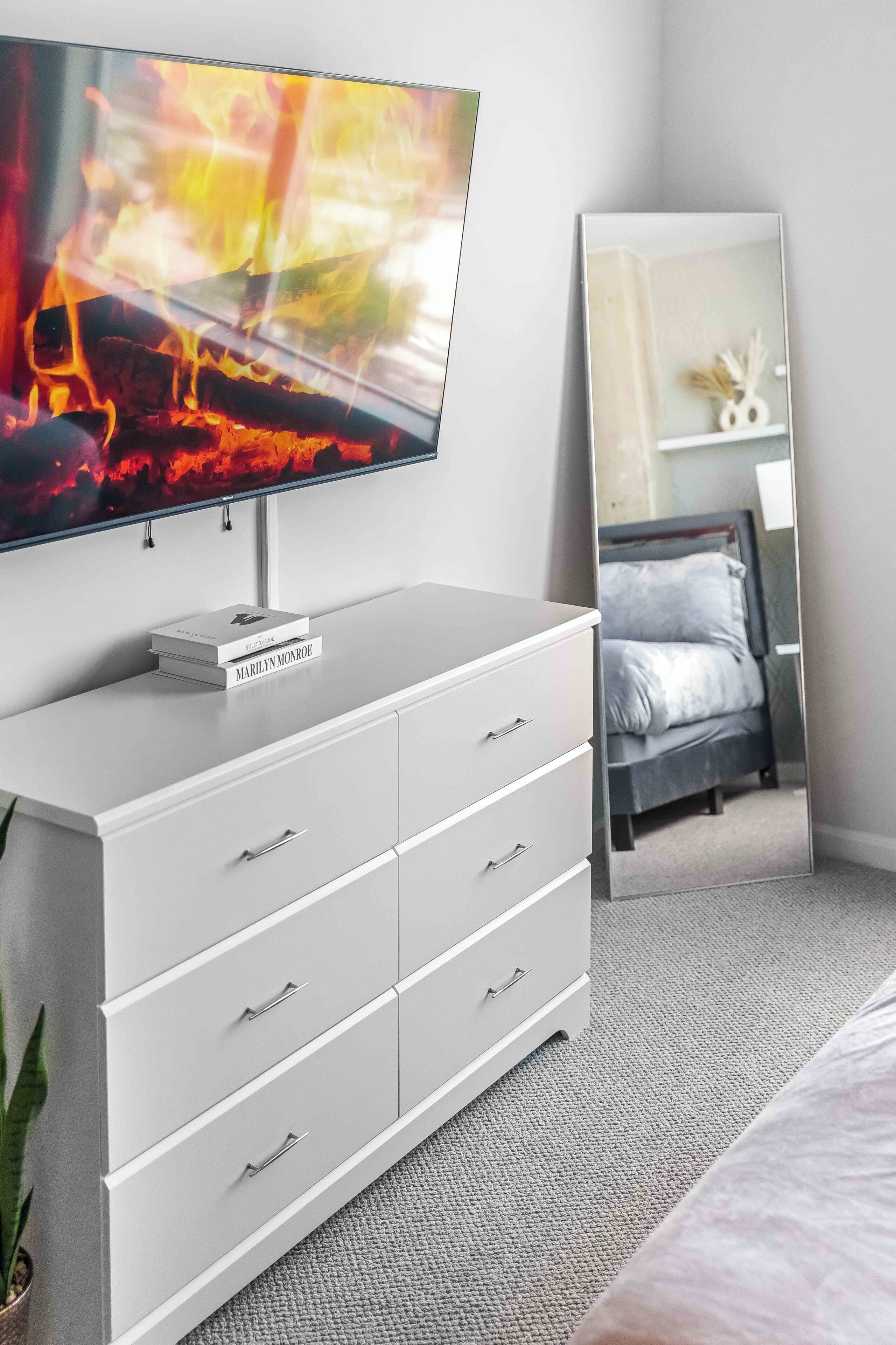 Un dormitorio con una televisión montada en un tocador blanco, una cama con cabecera gris y un espejo grande que refleja la habitación.