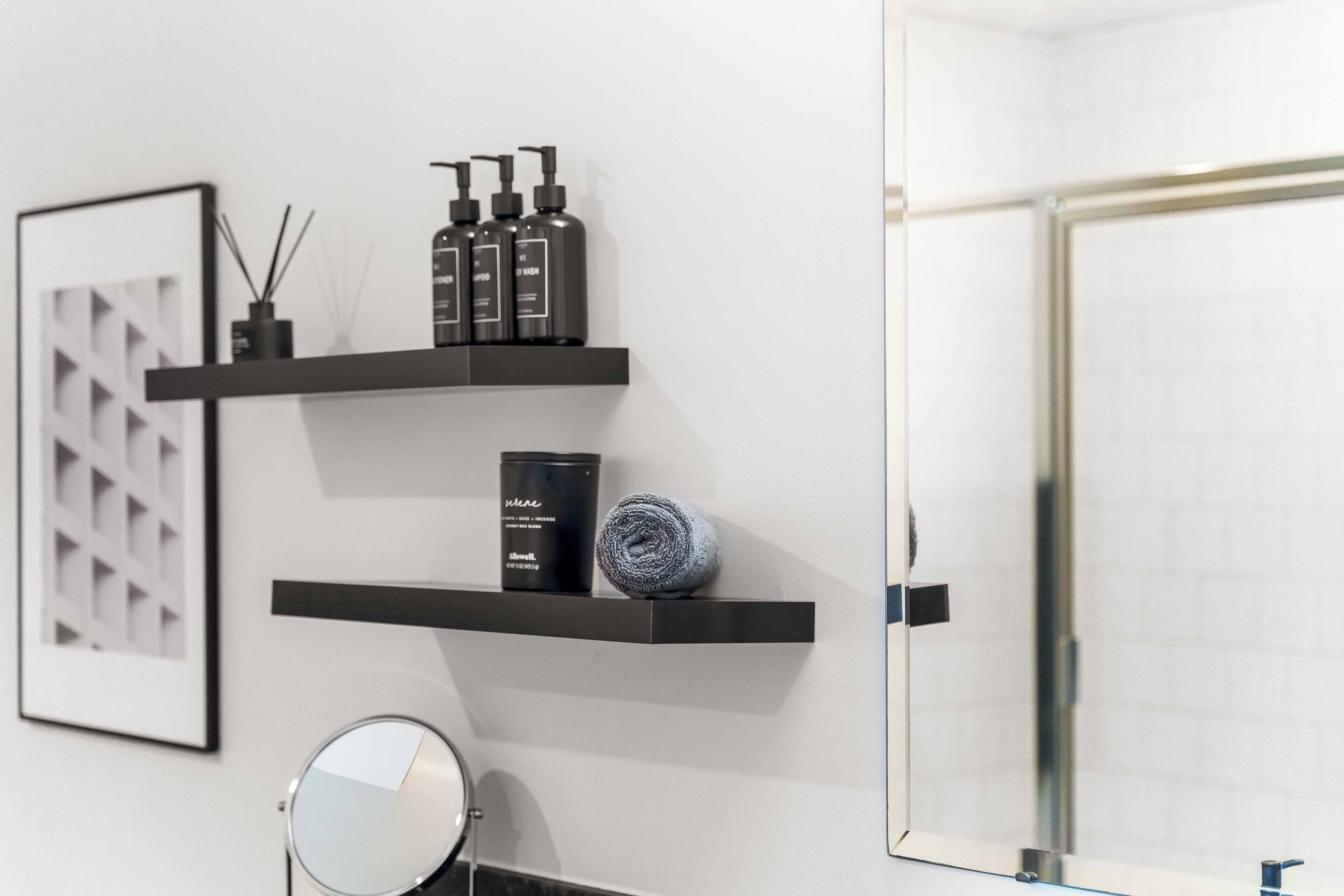 Une salle de bain avec trois étagères noires où sont rangés les articles de toilette, un miroir rond et un tableau encadré accroché au mur.
