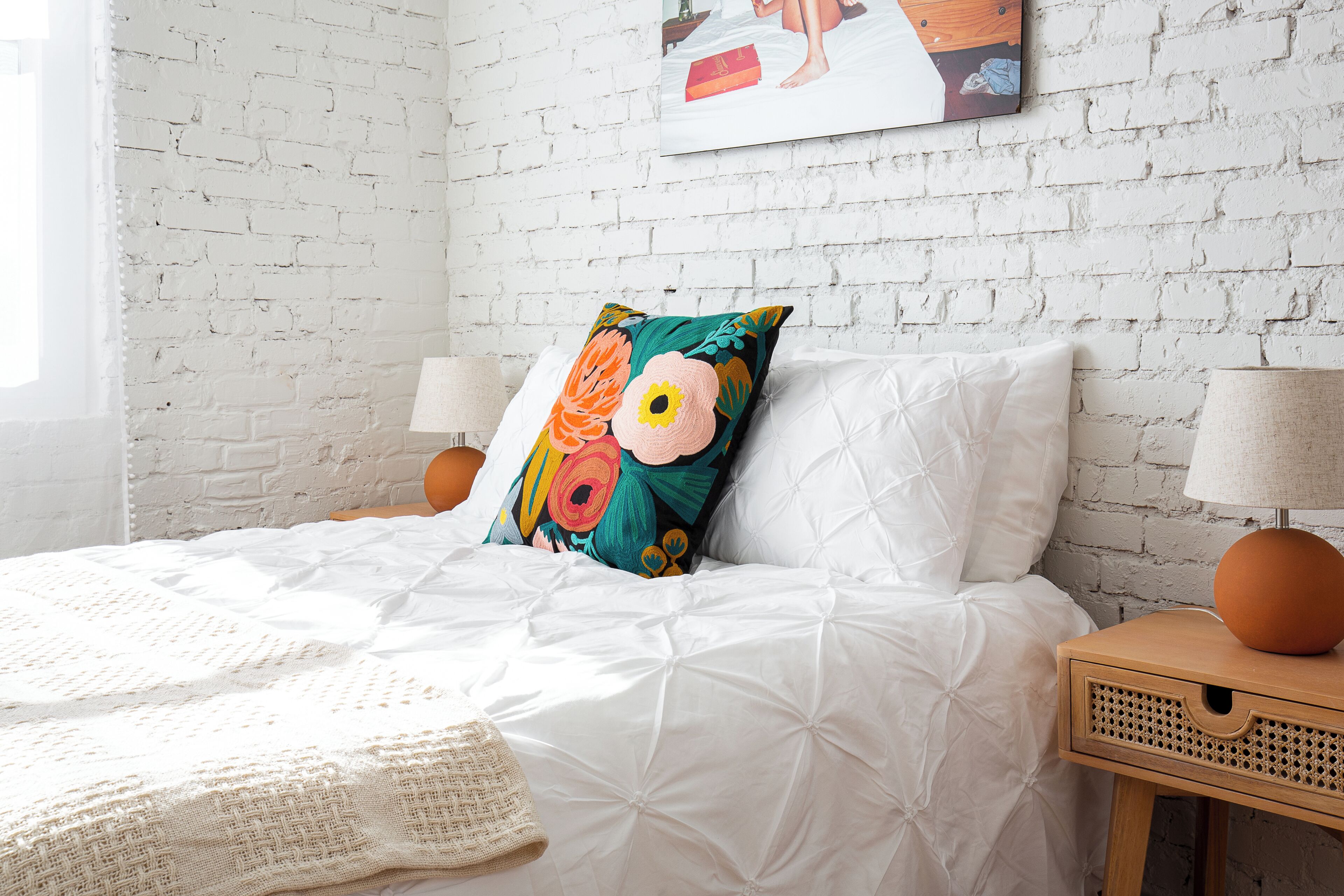 Een net opgemaakt bed met witte lakens en een kleurrijk kussen, een bedlampje en een klein houten nachtkastje met een lamp.