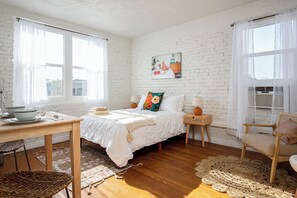 Een slaapkamer met een witte baksteenmuur, een bed met witte beddengoed, een houten tafel met een kom en een lepel, een rieten stoel en een geweven vloerkleed.