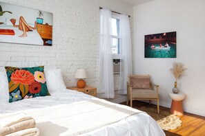 Een slaapkamer met een witte bakstenen muur, een bed met wit beddengoed, een stoel, een nachtkastje met een lamp en een schilderij van een boot op het water.