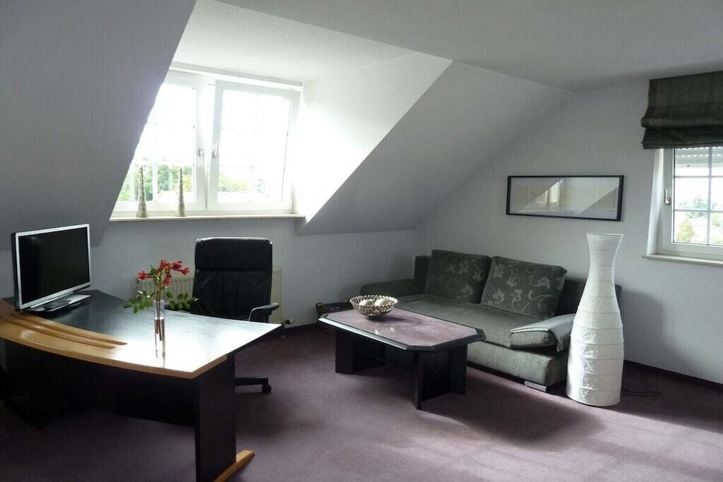 Una habitación con escritorio, una silla, sofá, un jarrón grande y ventana con vistas.