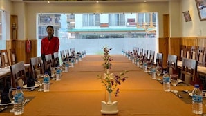 Un salón de conferencias con una mesa larga, sillas, botellas de agua y un jarrón con flores.