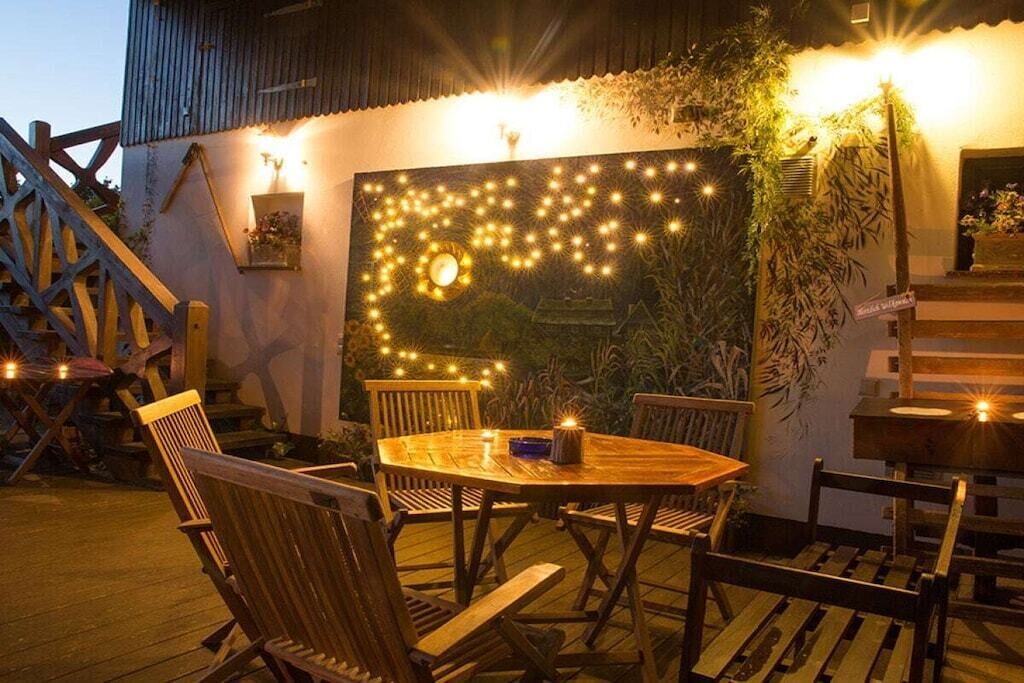 An outdoor dining area with a wooden table and chairs, surrounded by a wall decorated with string lights and plants.