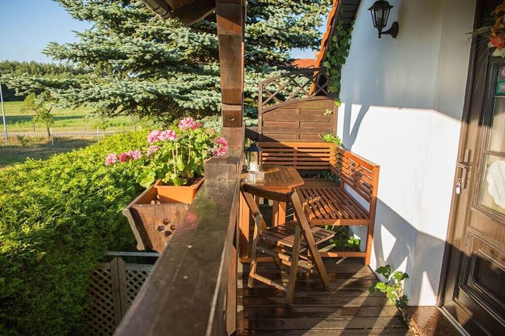 Una terrassa de fusta amb una banca i taula, envoltada de vegetació i un edifici blanc.