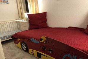 Quarto com colcha vermelha com estampa de carro, mesa de cabeceira branca e janela com cortinas.