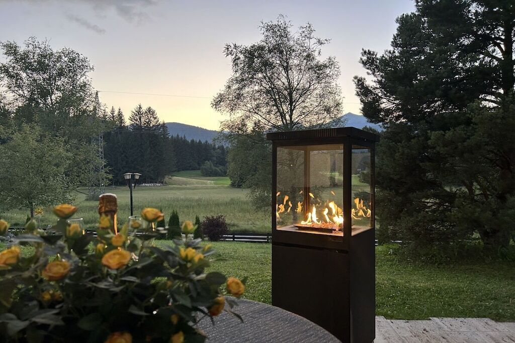 Ein im Freien stehender, gläserner Kamin mit sichtbarem Feuer, umgeben von einem Garten mit gelben Blumen, vor einer Baumlandschaft und weiter Ferne.