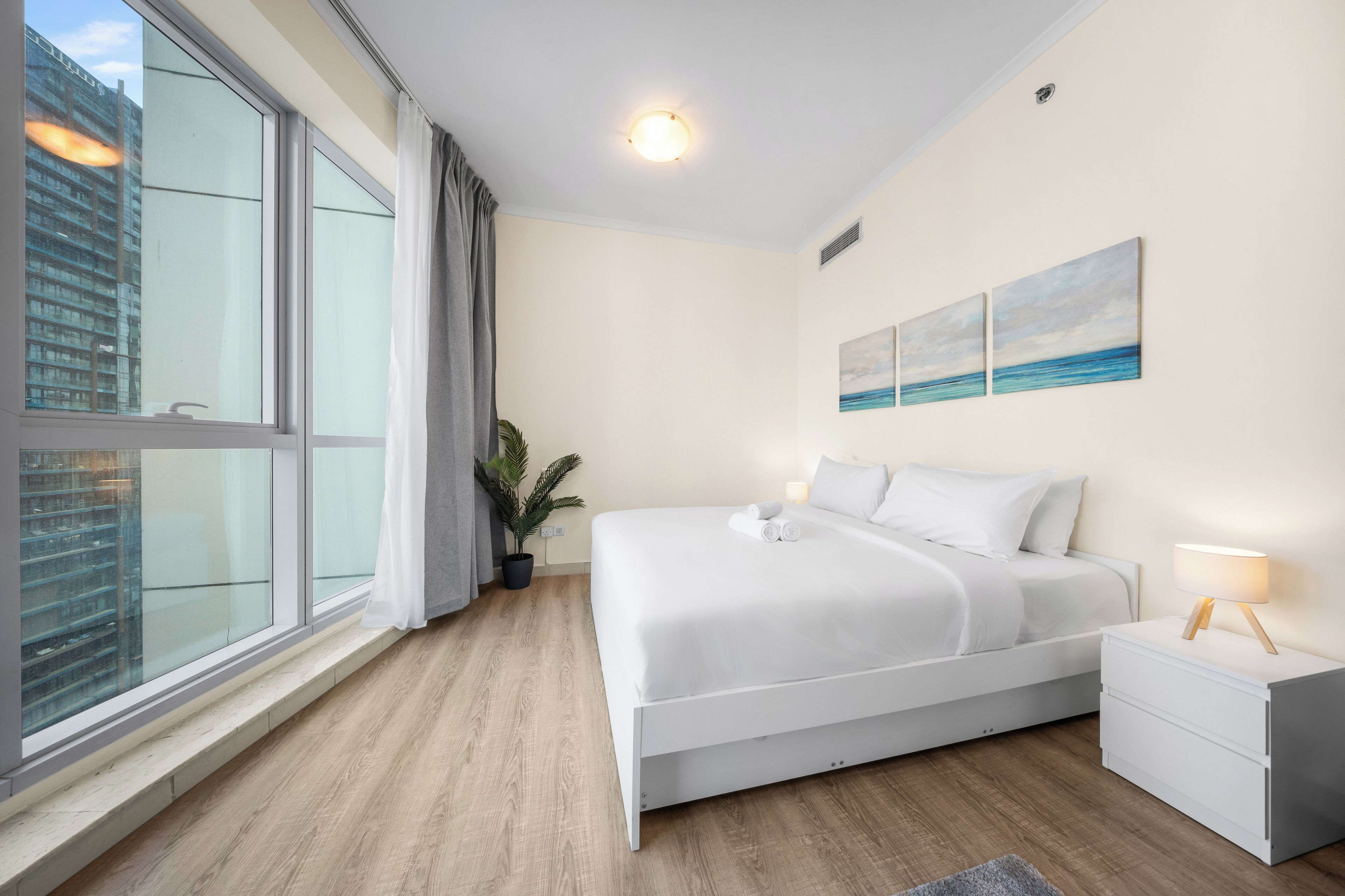 A modern hotel room with a large window, a bed with white linens, a nightstand, a potted plant, and a painting of a seascape.