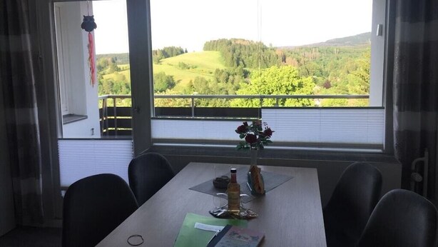 Uma área de refeições com mesa posta para dois, janela com vista para uma paisagem verde e um adorno pendurado.