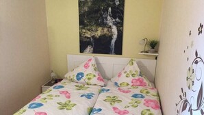 Quarto com cama, mesa de cabeceira, prateleira na parede com uma planta e pintura de uma cascata.