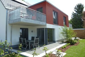Ein modernes, zweistöckiges Haus mit einem Balkon, einer Terrasse mit Tisch und Stühlen und einem gepflegten Garten.