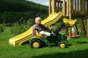 A child on a green toy tractor with a yellow slide in a grassy outdoor area. - Schulte Holiday Farm (Schmallenberg)