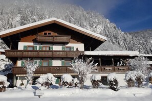 A multi-story wooden chalet with balconies, surrounded by snow-covered trees and a mountain backdrop. - Holiday apartment with 1 bedroom (Reit im Winkl)