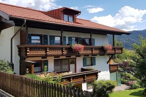 Een gebouw met twee verdiepingen, een balkon, een houten omheining en een bergachtig landschap op de achtergrond.