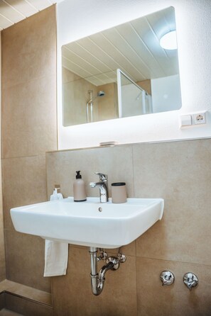 Un baño con un lavamanos blanco, un espejo encima y una ducha que se ve en el reflejo.