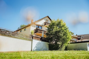 Een woonhuis met twee verdiepingen, een balkon en een boom in de voortuin, onder een heldere lucht.