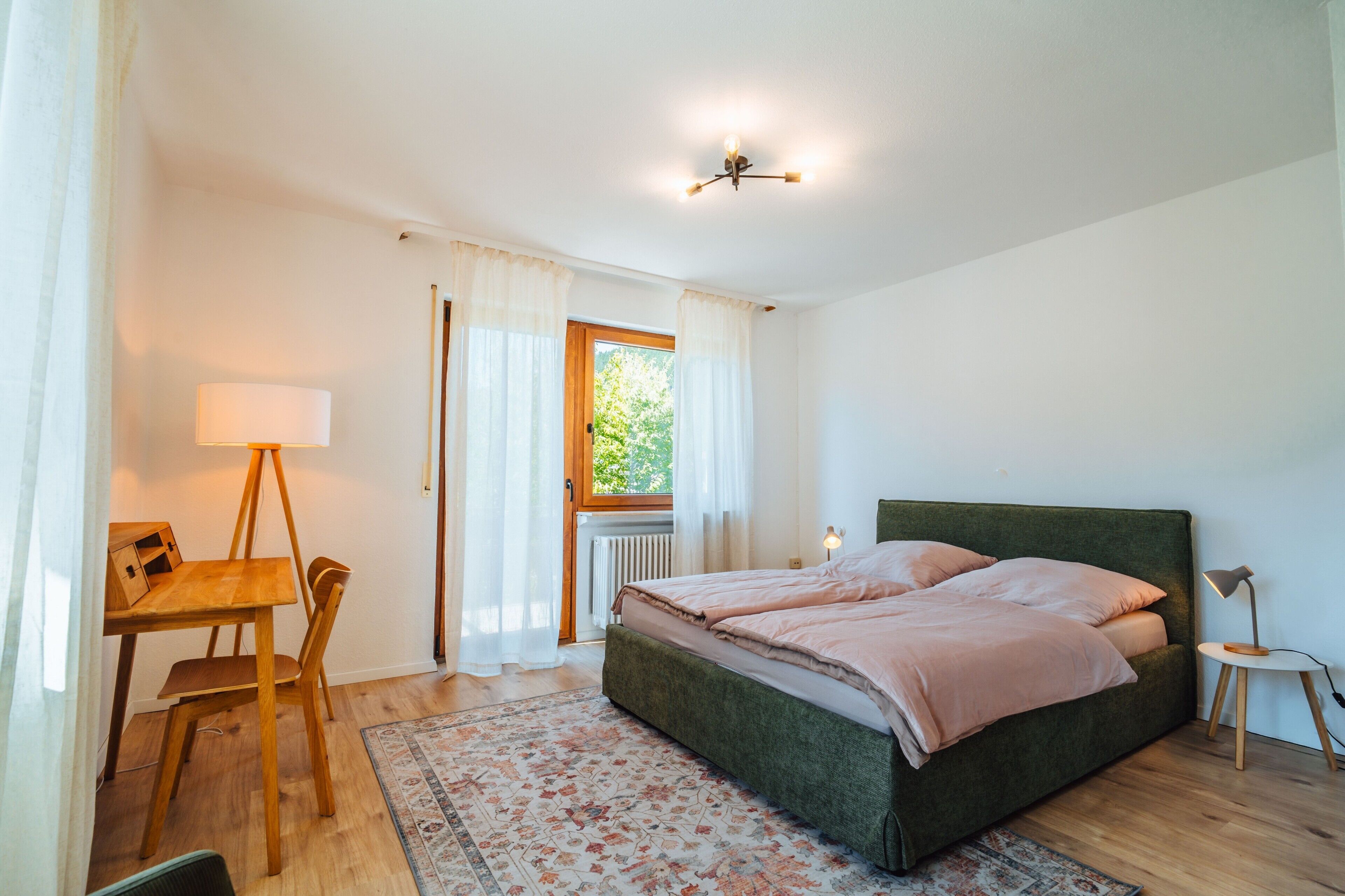 Ein Schlafzimmer mit einem Bett, einem Schreibtisch, einem Stuhl, einer Lampe und einem Fenster mit Vorhängen.