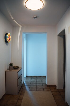 Een gang met een plafondlamp, een decoratief object aan de muur, een kleine tafel met een plant en een deur naar een andere kamer.