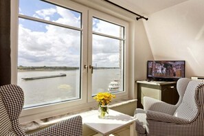 A room with a large window overlooking a body of water, a television on a cabinet, and a table with a vase of flowers. - "Ferryman" in the cottage on the Rhine (Rees)