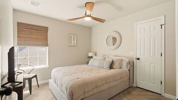 A bedroom with a bed, a chair, a window with blinds, a ceiling fan, a door, and a wall-mounted mirror.