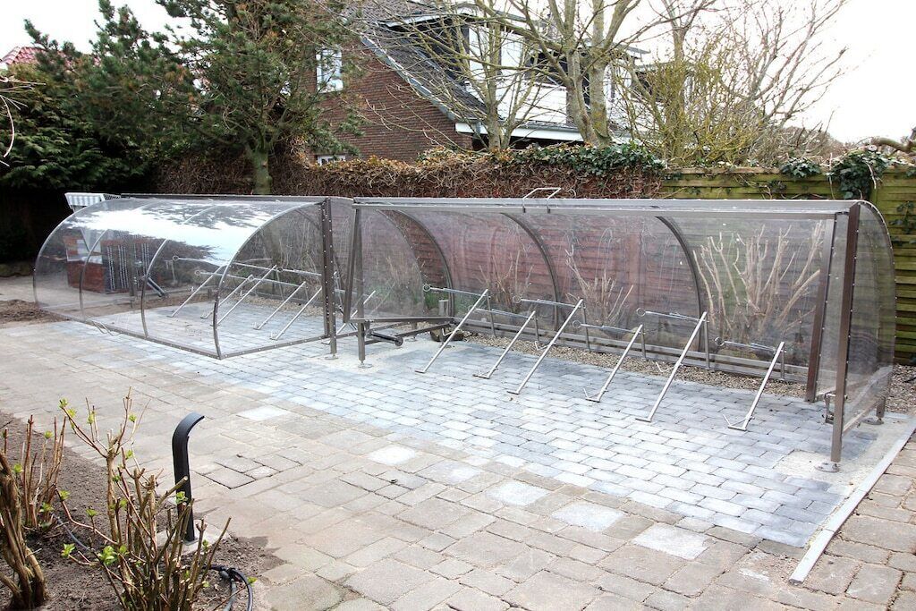 Ein überdachter Außenbereich mit Metallgestellen und einem transparenten Dach, umgeben von einer Pflasterterrasse.