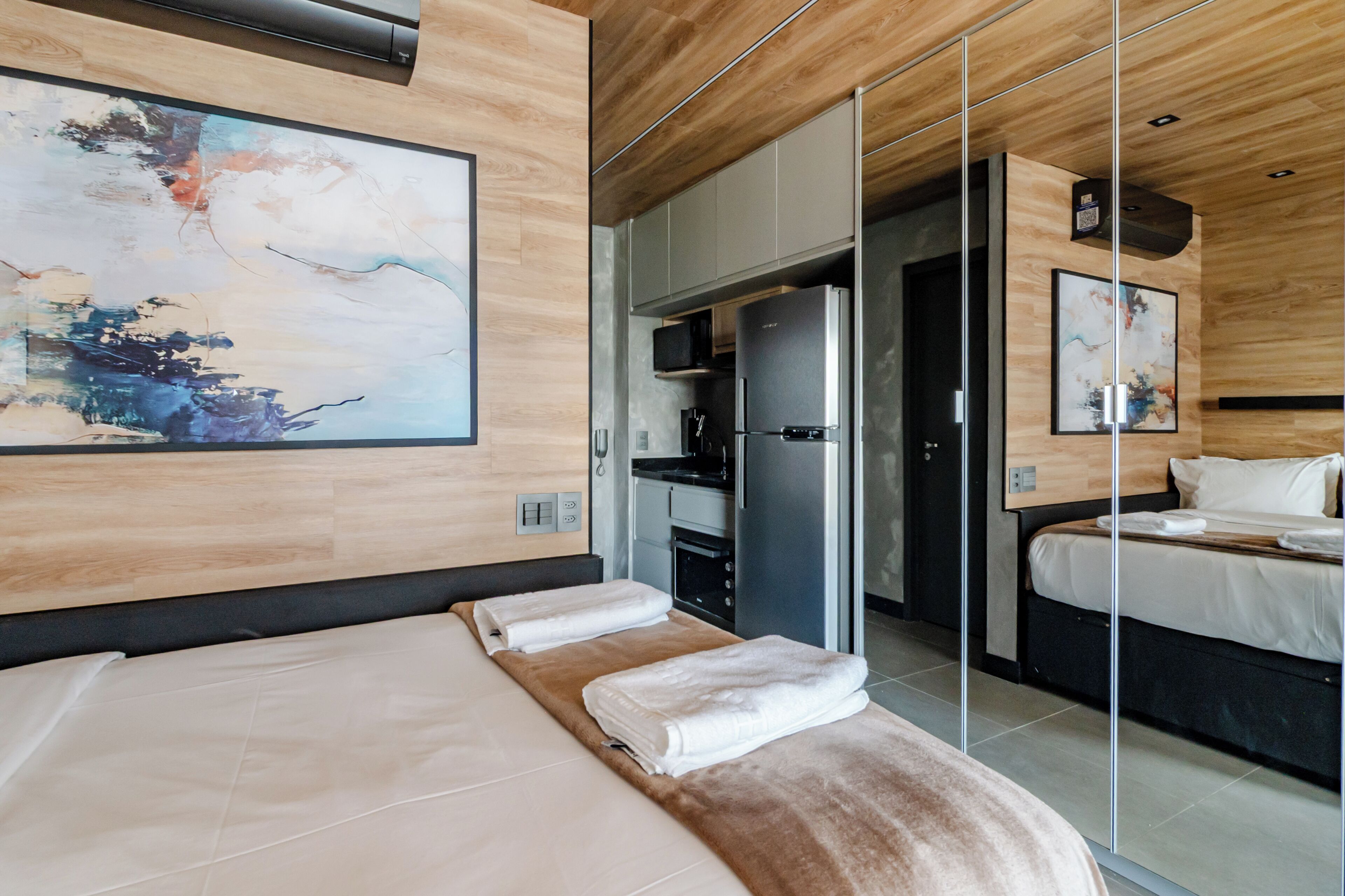 Une chambre d’hôtel moderne dotée d’un grand lit, d’une armoire intégrée, d’une kitchenette et d’un grand miroir qui reflète la pièce.