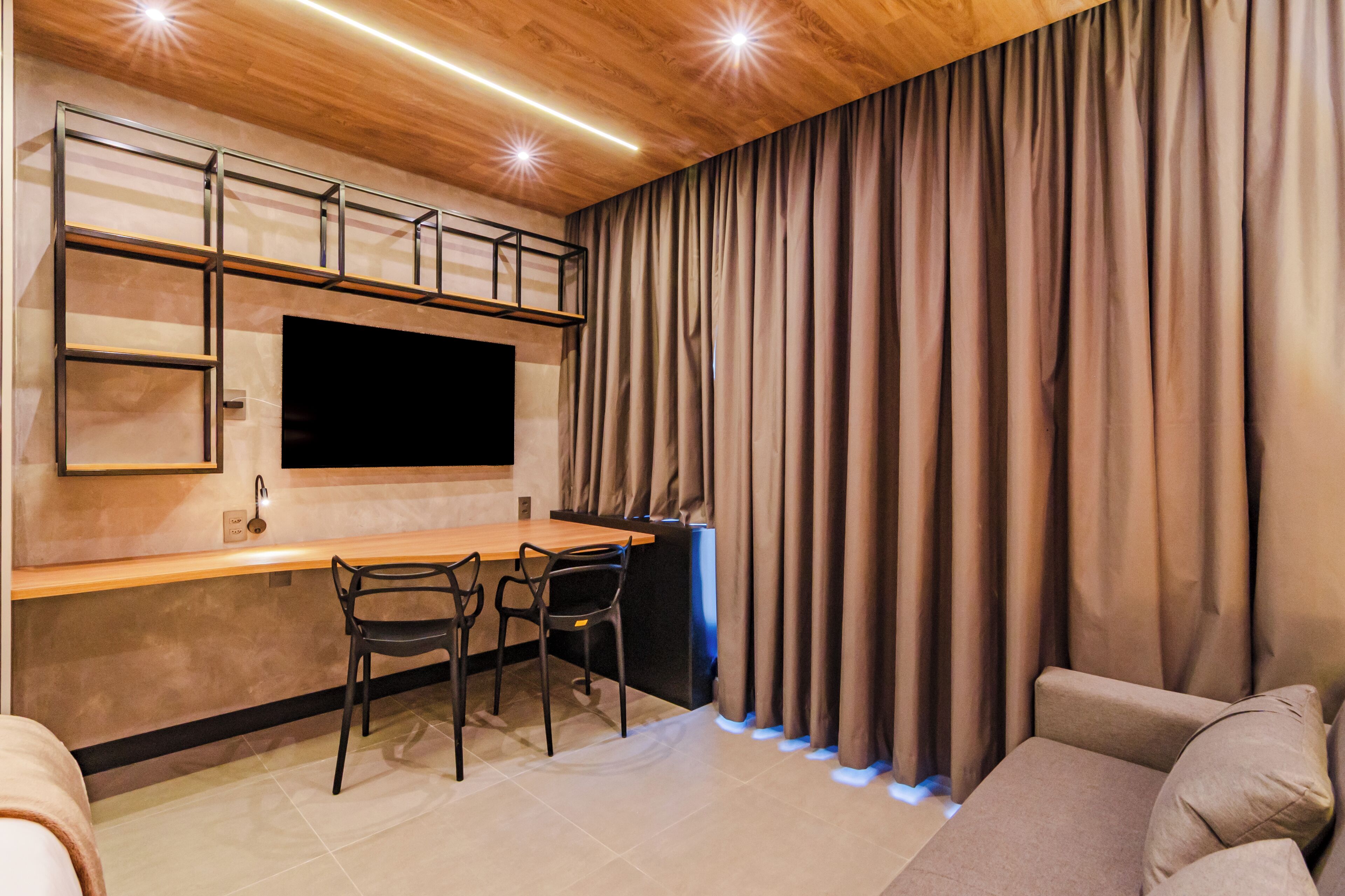 Ein moderner Wohnraum mit einem an der Wand befestigten Flachbildfernseher, einem Holztisch, zwei Stühlen, einem Sofa und einem großen Fenster mit Vorhängen.