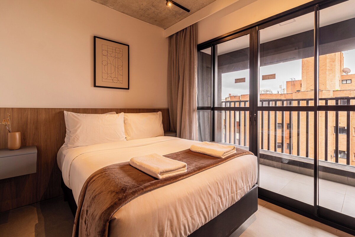 Camera d'albergo con un letto grande, un comodino, un'opera d'arte incorniciata sulla parete e vista sulla città attraverso un'ampia finestra.
