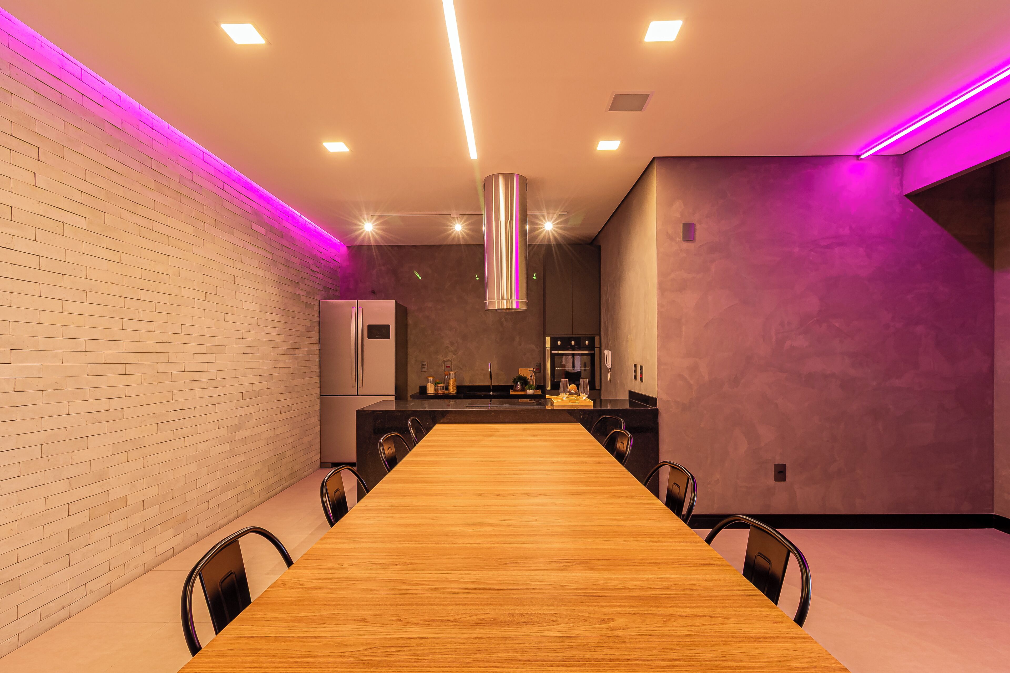 Un espace de restauration moderne avec une longue table en bois, des chaises, une cuisine en arrière-plan et un éclairage d’ambiance violet.