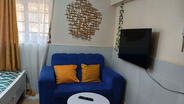 A room with a blue sofa, a white table, a TV mounted on the wall, and a bed with a patterned bedspread. - Studio airbnb mirema drive (Nairobi)